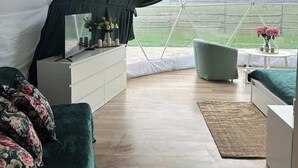 A tented room with a bed, a sofa, a side table, and a view of a green field.