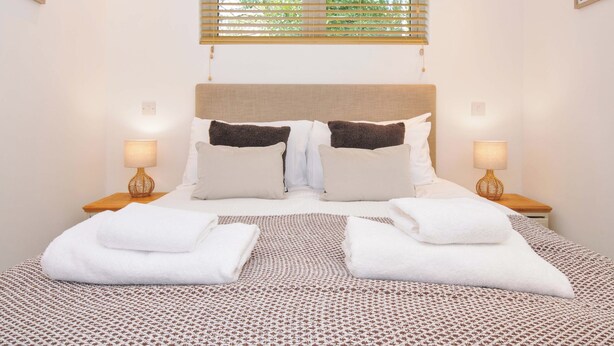 A neatly made bed with white and brown bedding, two bedside lamps, and a window with blinds. - 3 Faraway Fields (Liskeard)