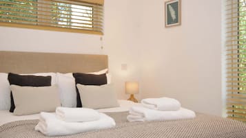 Una cama bien hecha con ropa de cama blanca y marrón, una lámpara de noche y una impresión botánica enmarcada en la pared.
