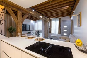 A modern kitchen with a black induction cooktop, wooden beams, and a dining area with a sofa and a yellow fruit bowl. - Le Caveau Dijonnais – Exceptional Duplex (Dijon)