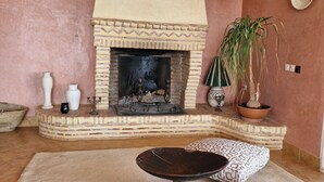 Een woonkamer met een open haard, een ronde salontafel, gekleurde kussens, een potplant en decoratieve vazen.