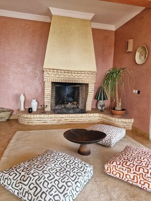 A living room with a fireplace, a round coffee table, patterned cushions, a potted plant, and decorative vases.