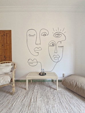 A room with a wall mural, a wooden table with bottles, a wicker chair, and a bed.