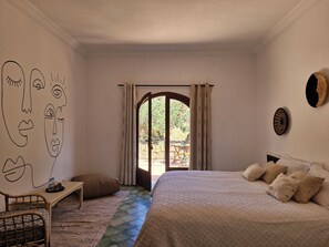 A bedroom with a bed, a chair, a small table, a wall with a drawing, and a view through an open door.