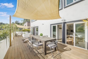 Eine Holzterrasse mit Tisch und Stühlen, ein großes Sonnensegel und Blick auf das Haus.