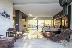 A modern living room with a sofa, armchair, and a large window with a view of greenery. - Bowmont Glass Estate - Sleek, Architectural and Unique Contemporary Estate (Beverly Hills)