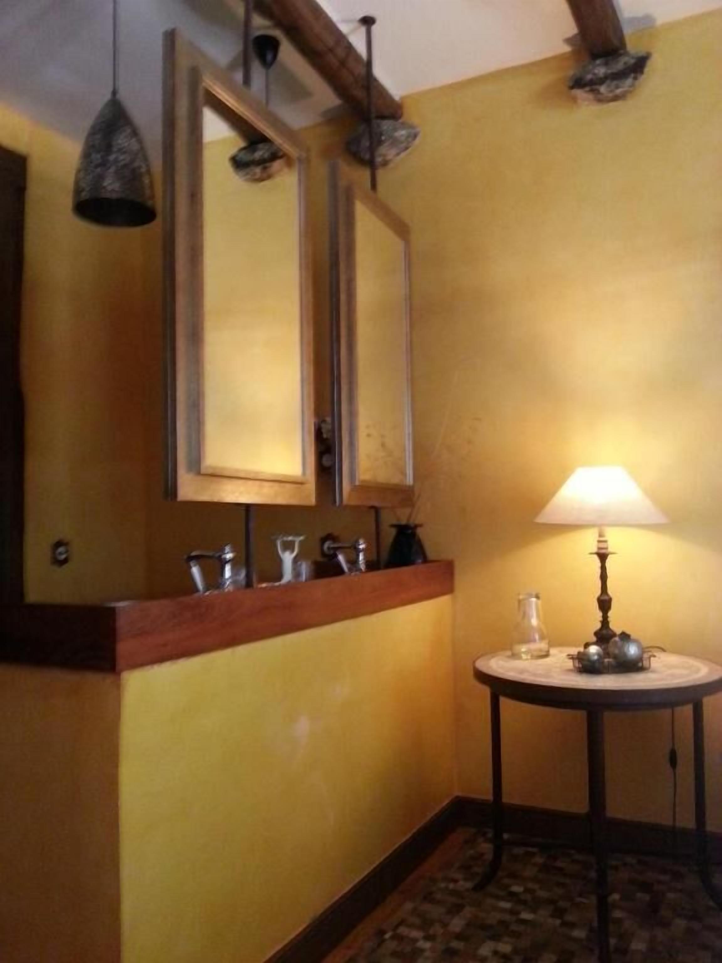 A bathroom with a wooden vanity, a mirror, a sink, a round table with a lamp, and a patterned floor.