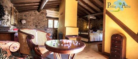 A rustic room with a stone wall, wooden ceiling, a bed, a table with chess pieces, and a sign reading 'Peña de Alba'.