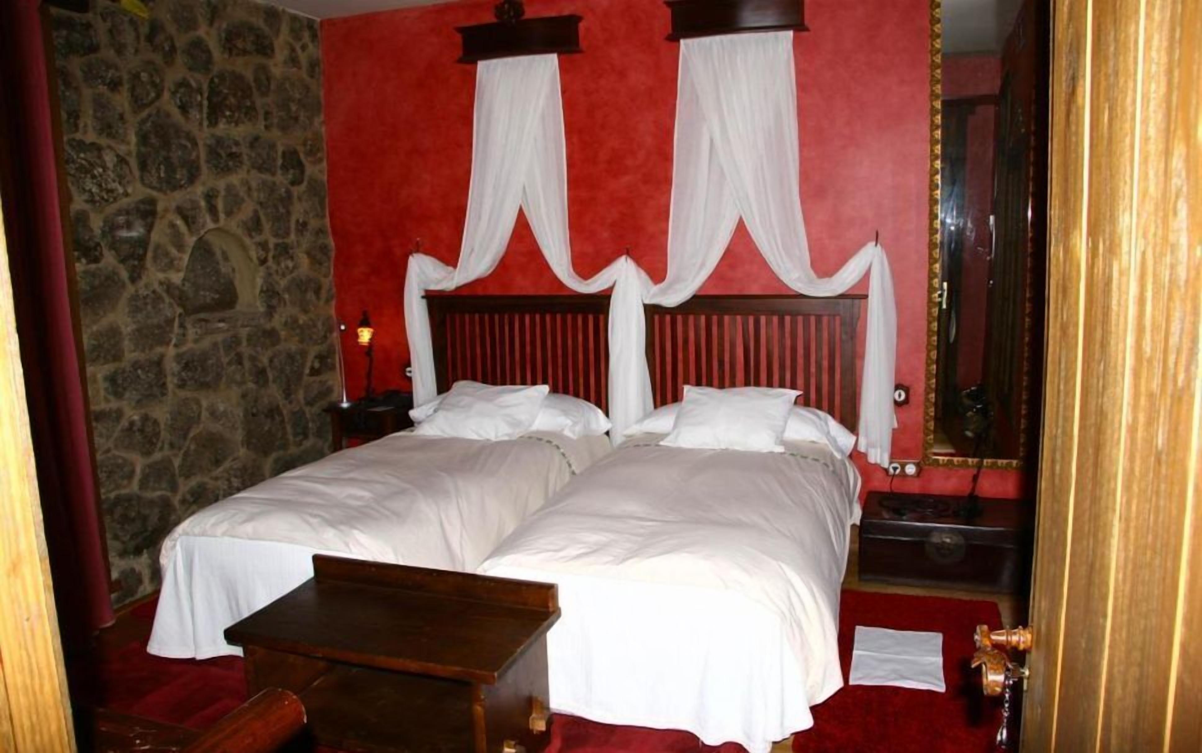 A double bed with white linens, a wooden nightstand, a stone wall, and a red carpeted floor.