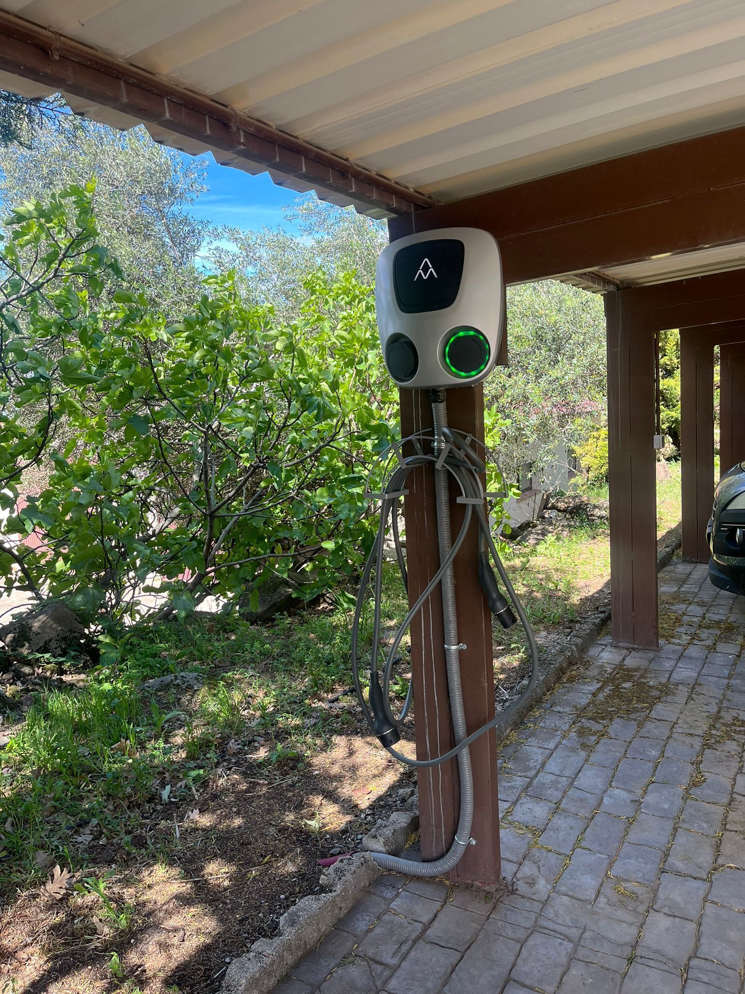Une borne de recharge pour véhicules électriques installée en extérieur, dotée d’un poteau en bois, de câbles et d’un indicateur lumineux vert.