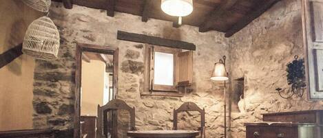A rustic room with stone walls, wooden beams, a round table with chairs, a hanging light fixture, and a floor lamp.