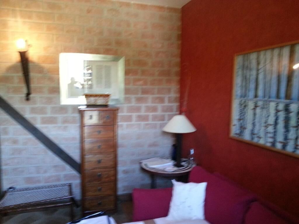A room with a brick wall, a wooden dresser, a red couch, a small table with a lamp, and a framed picture on the wall.