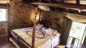 Une chambre à coucher rustique avec un lit à baldaquin, des murs en pierre, un plancher en bois et une fenêtre offrant une vue agréable.