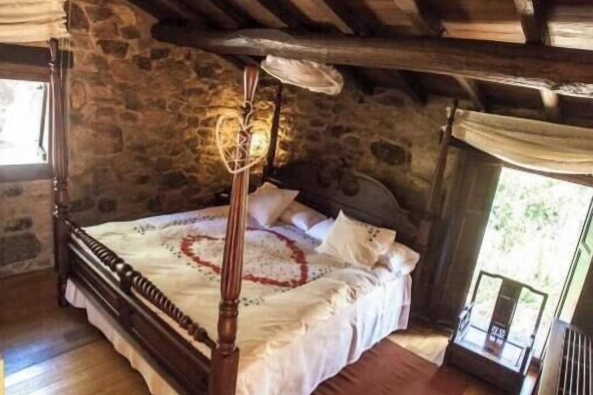 A rustic bedroom with a four-poster bed, stone walls, wooden flooring, and a window with a view.