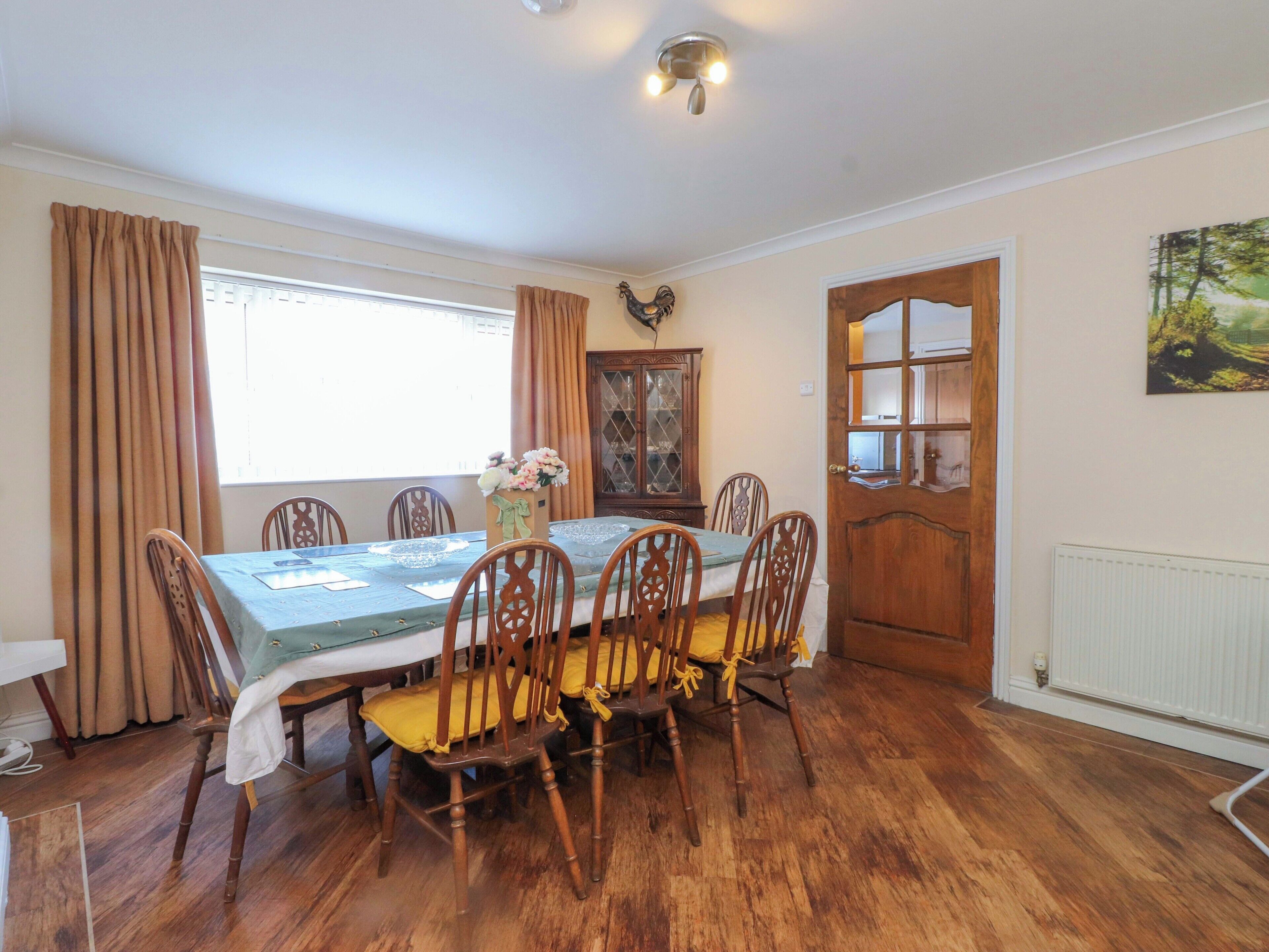 Uma sala de jantar com uma mesa posta para seis lugares, cadeiras de madeira, um armário e uma porta que dá para outro cômodo.