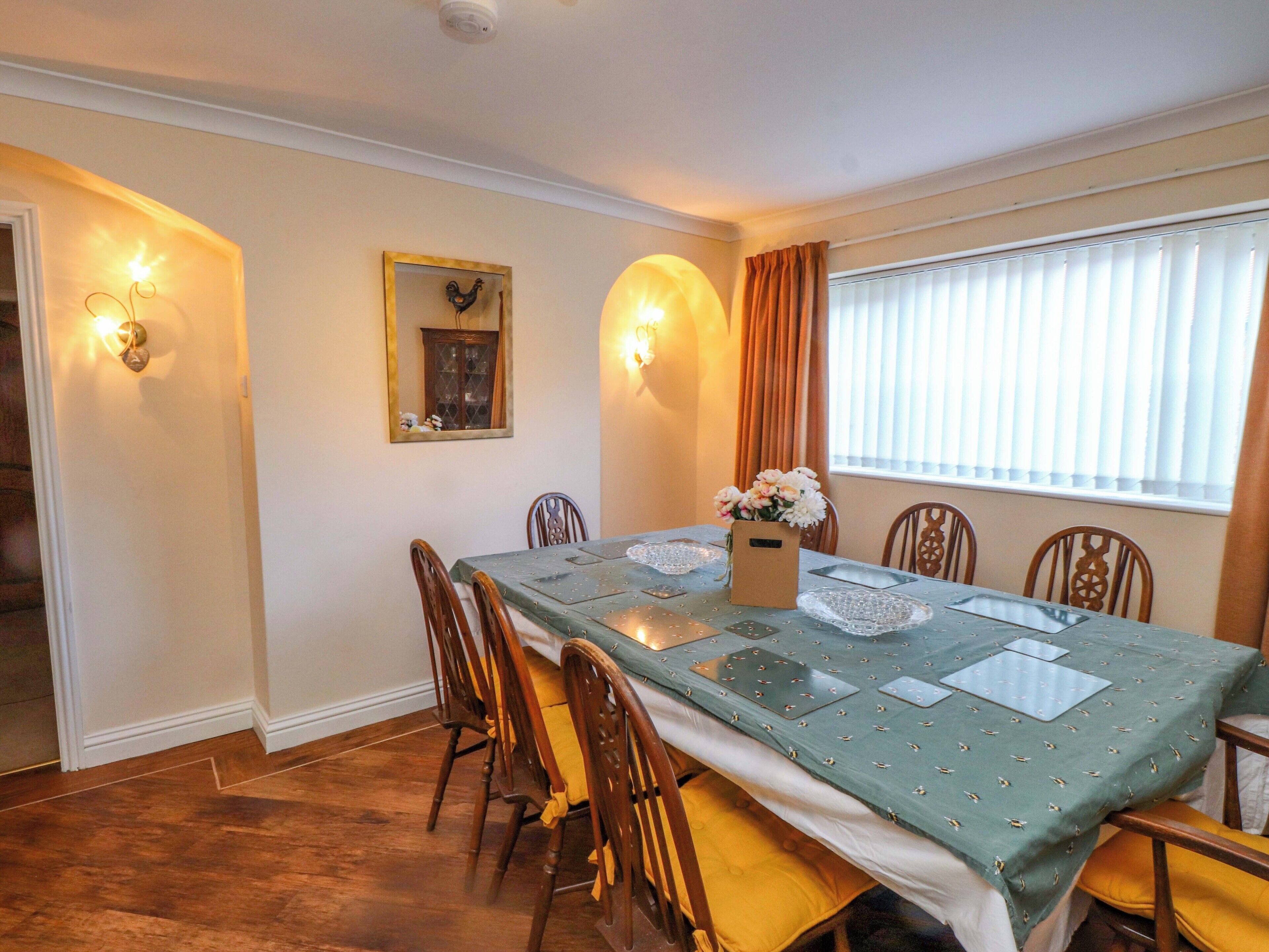 A dining room with a table set for six, surrounded by wooden chairs, a vase of flowers, and wall-mounted lights.
