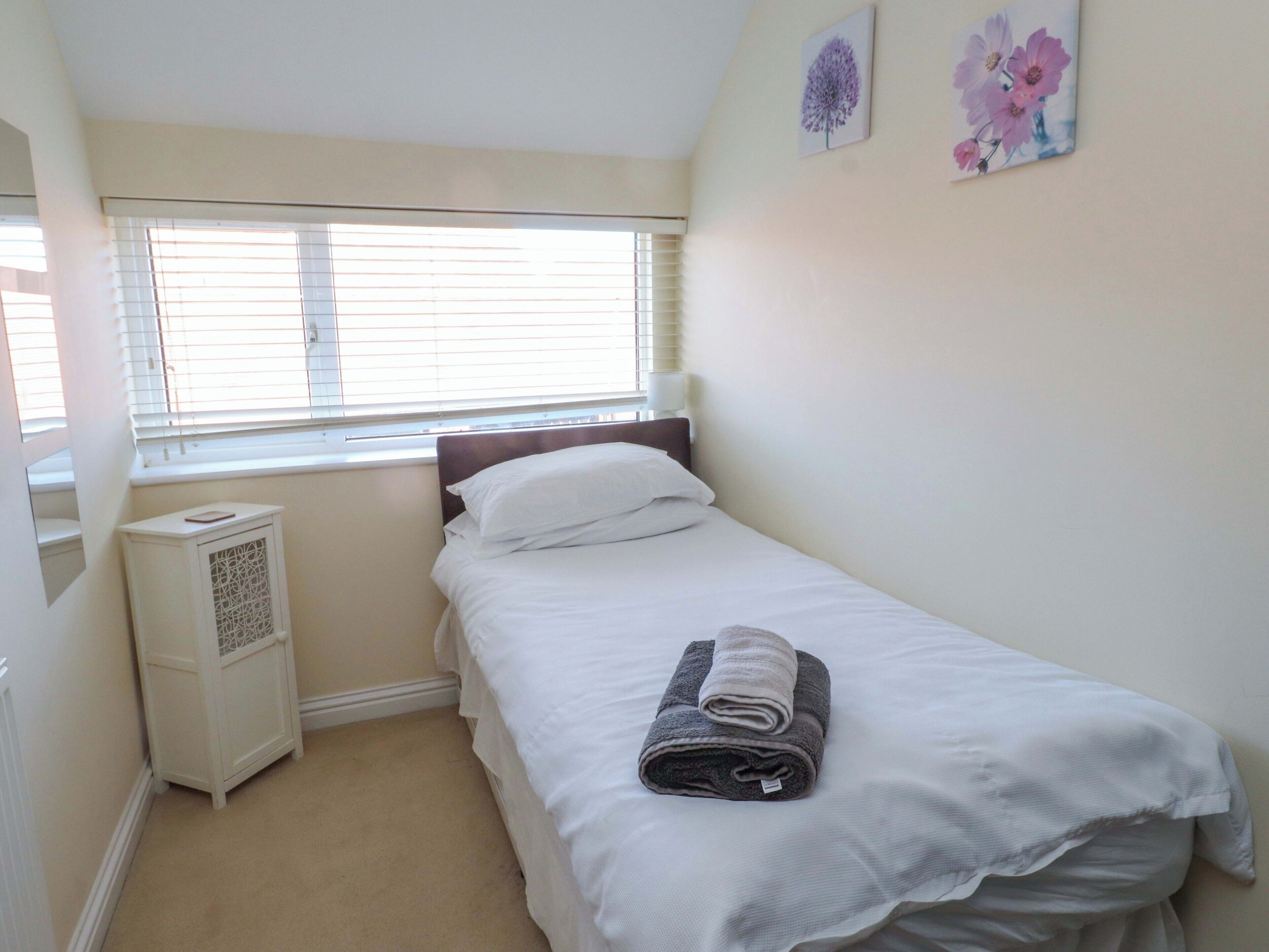 A small, neatly made bedroom with a single bed, a window with blinds, a radiator, and a small cabinet.