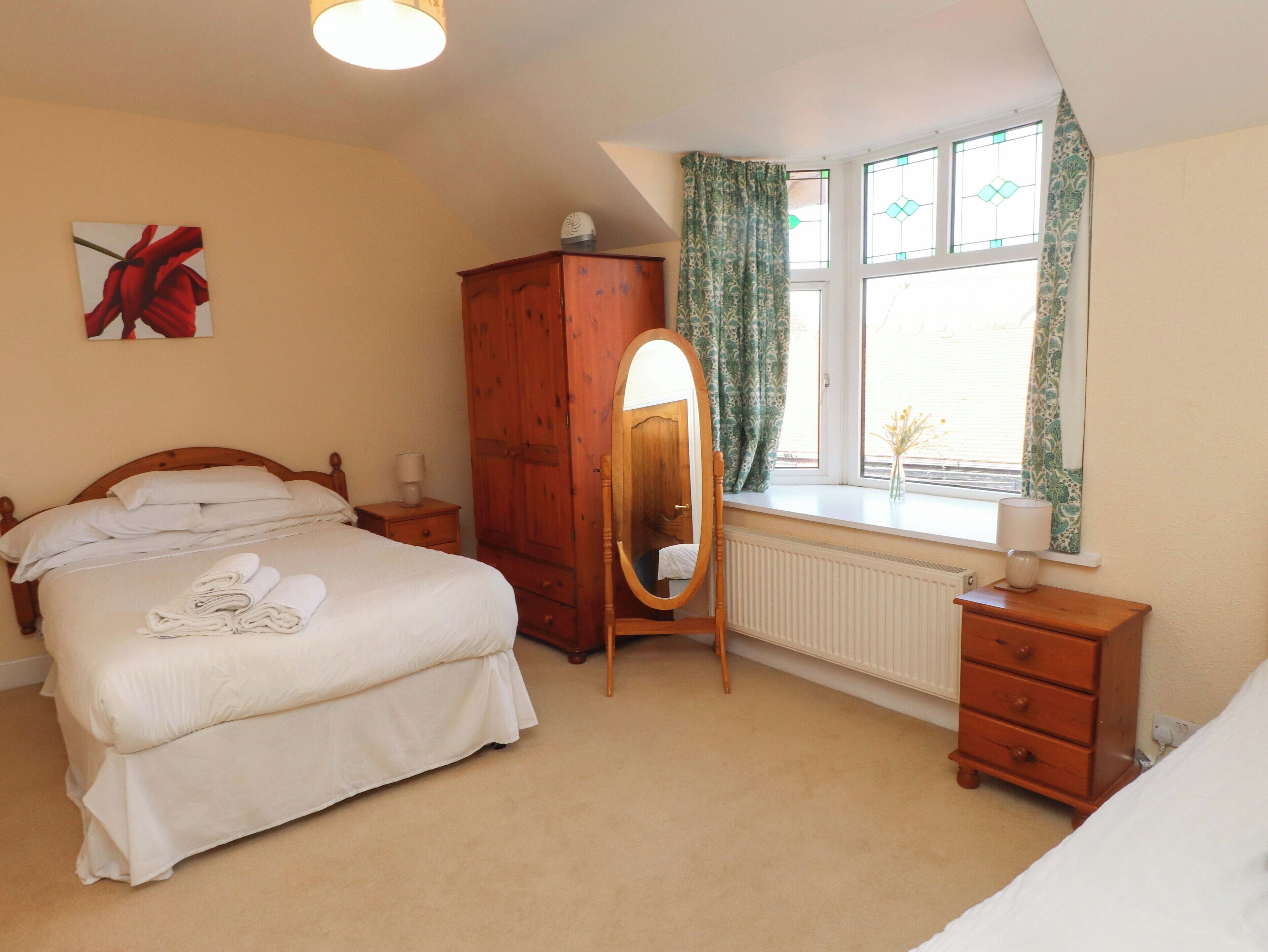 A bedroom with a bed, a wooden wardrobe, a dressing table, a mirror, and a window with curtains.