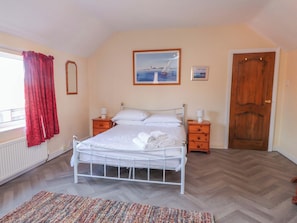 Un dormitorio con cama, dos mesitas de noche, una puerta de madera, un radiador y una ventana con cortinas rojas.