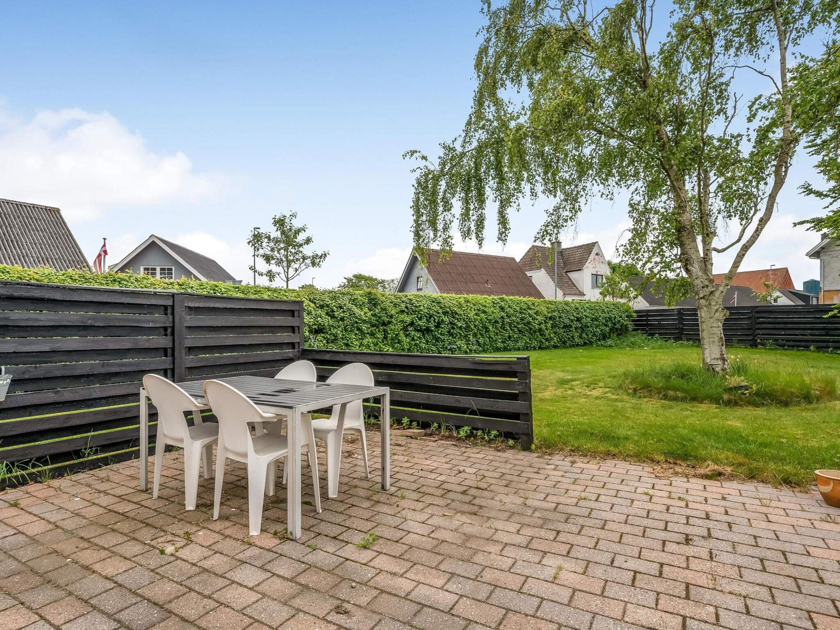 Eine Terrasse mit Tisch und Stühlen, ein gepflasterter Weg, ein Holzzaun und ein Heckenstreifen.