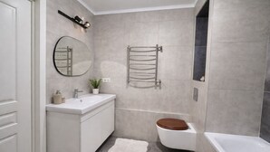 A modern bathroom with a white vanity, a round mirror, a towel rack, a toilet, and a bathtub.