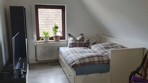 A bedroom with a bed, a radiator, a window with plants, and a TV stand.