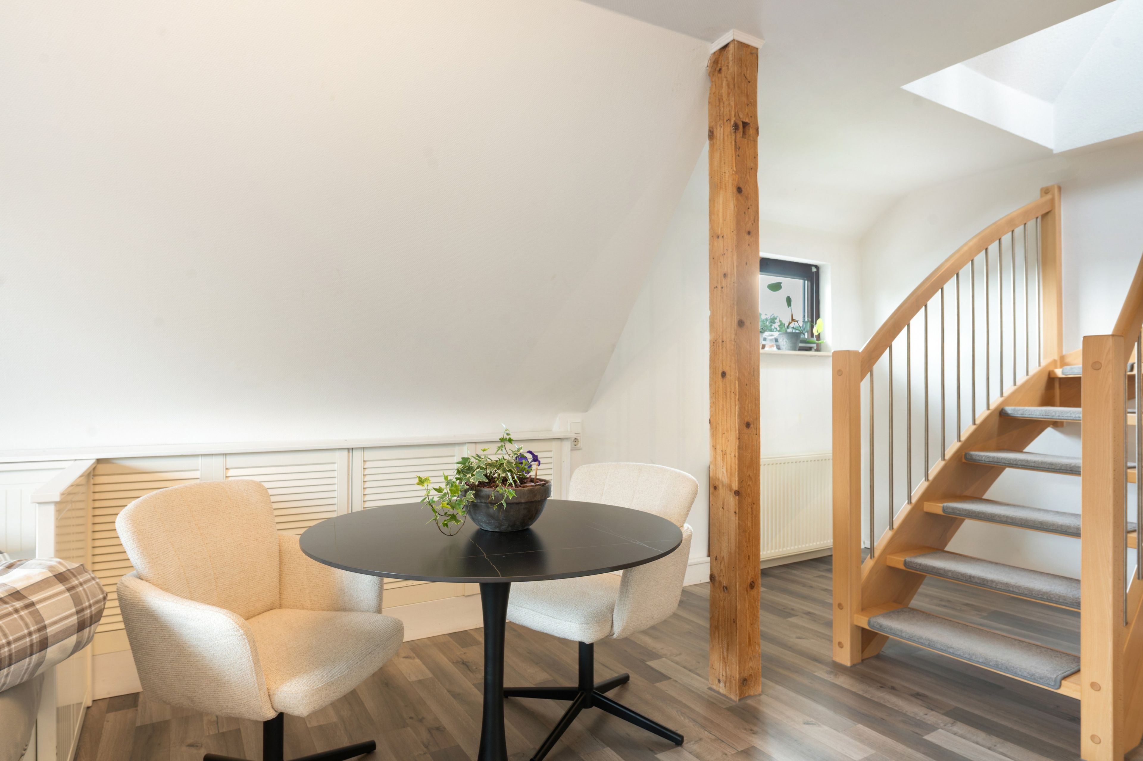 A modern loft bedroom with a skylight, a round table with a potted plant, a wooden staircase, and a view of the city.