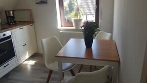 A kitchen with white cabinets, a wooden table, and chairs. There is a window with a view of trees and a potted plant on the windowsill.