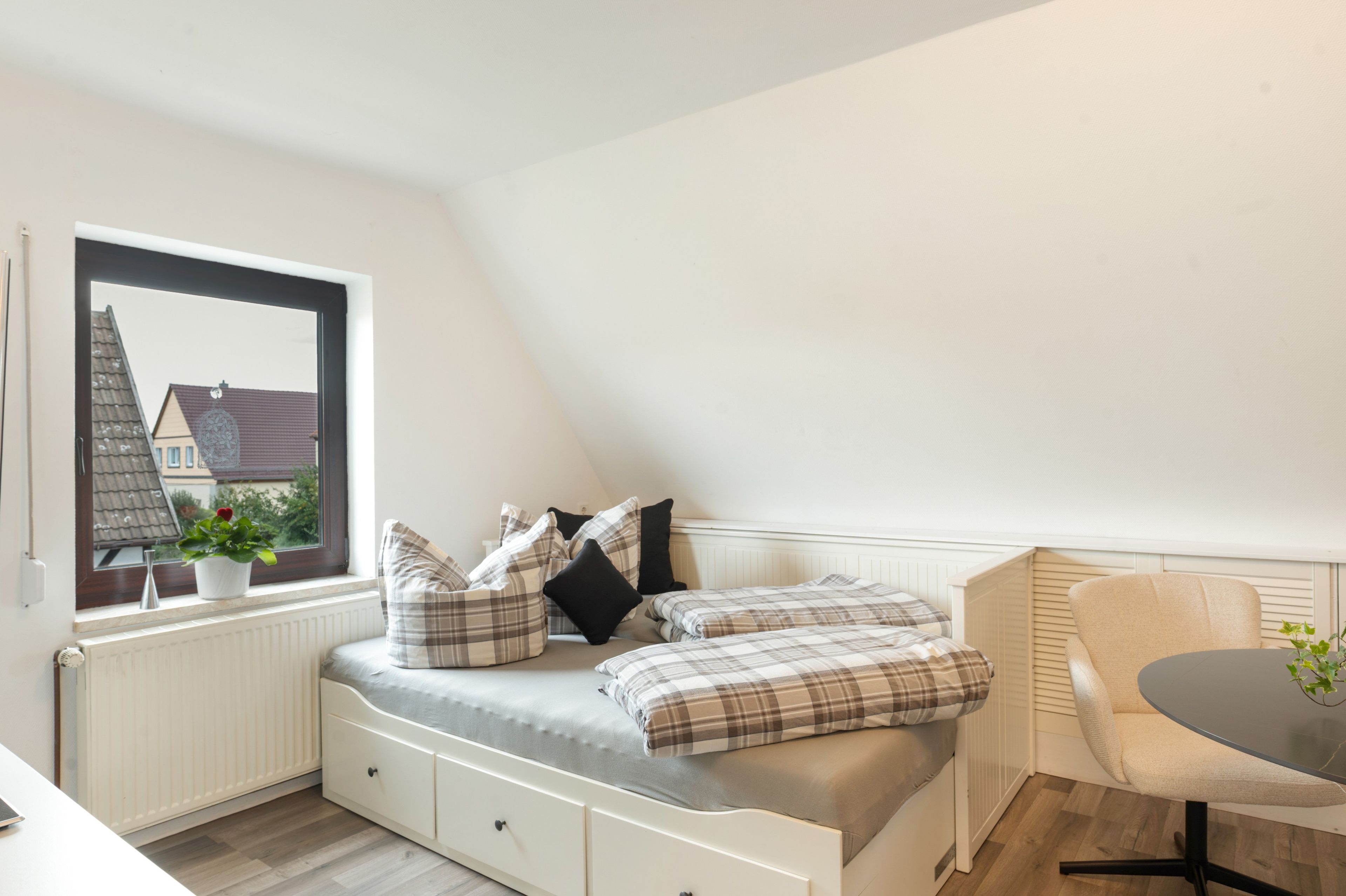 A small, cozy room with a bed, a chair, and a table. There is a window with a view of a house outside.