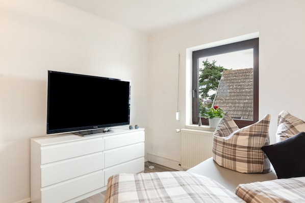 A bedroom with a television on a white dresser, a window with a view of a building and a plant, and a bed with plaid bedding.