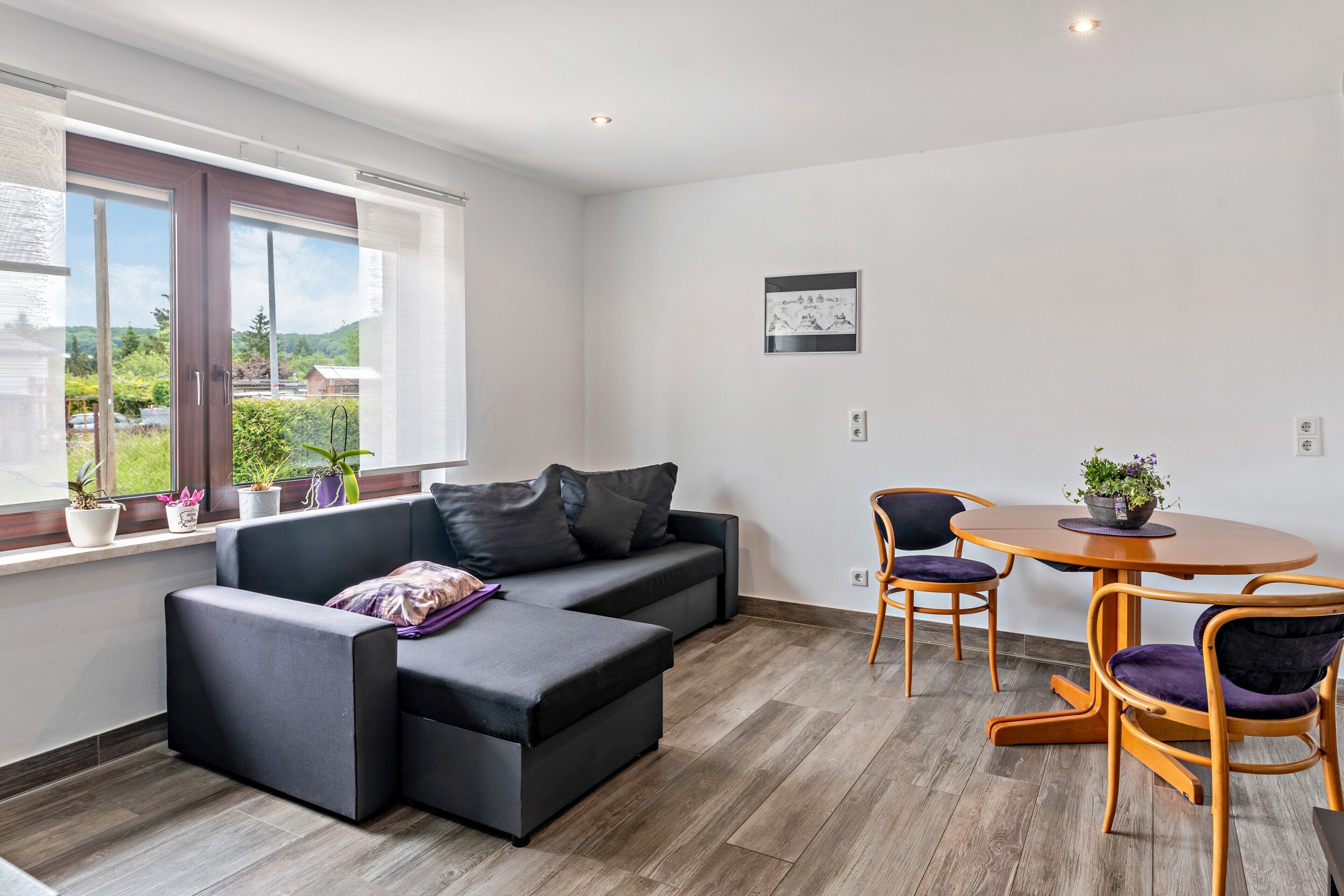 A modern living room with a sectional sofa, a dining table with chairs, and a view of greenery outside.