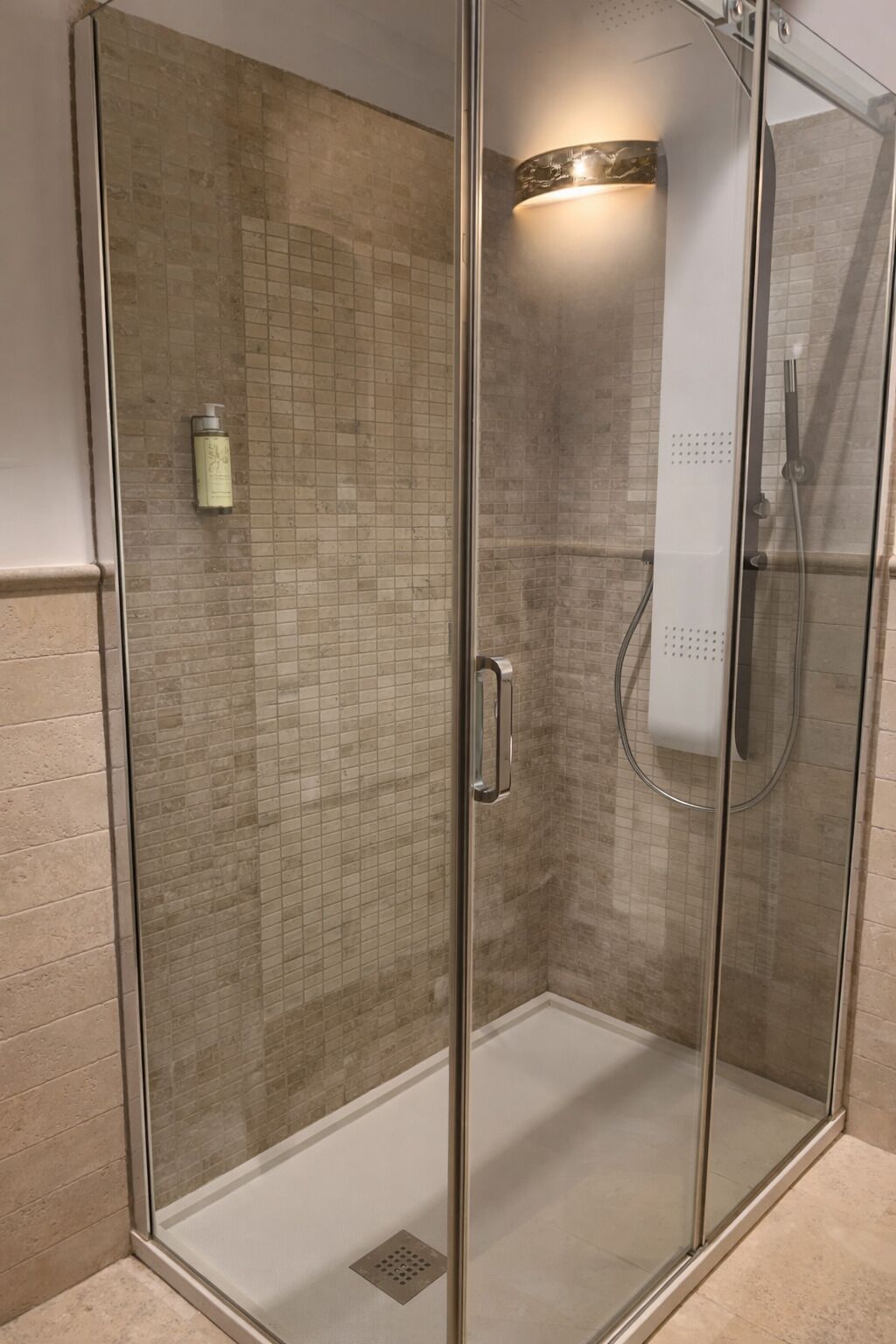 Lasiseinöillä varustettu suihkukaappi, jossa on laatoitettu seinä, käsisuihku ja seinällä oleva shampoopullo.