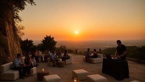 Un área de estar al aire libre con sofás y otomanas dispuestas para los huéspedes, una cabina de DJ y una vista panorámica del atardecer.