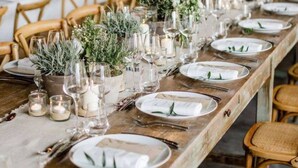 Una larga mesa de comedor dispuesta con platos blancos, cubiertos y cristalería, decorada con follaje y velas.