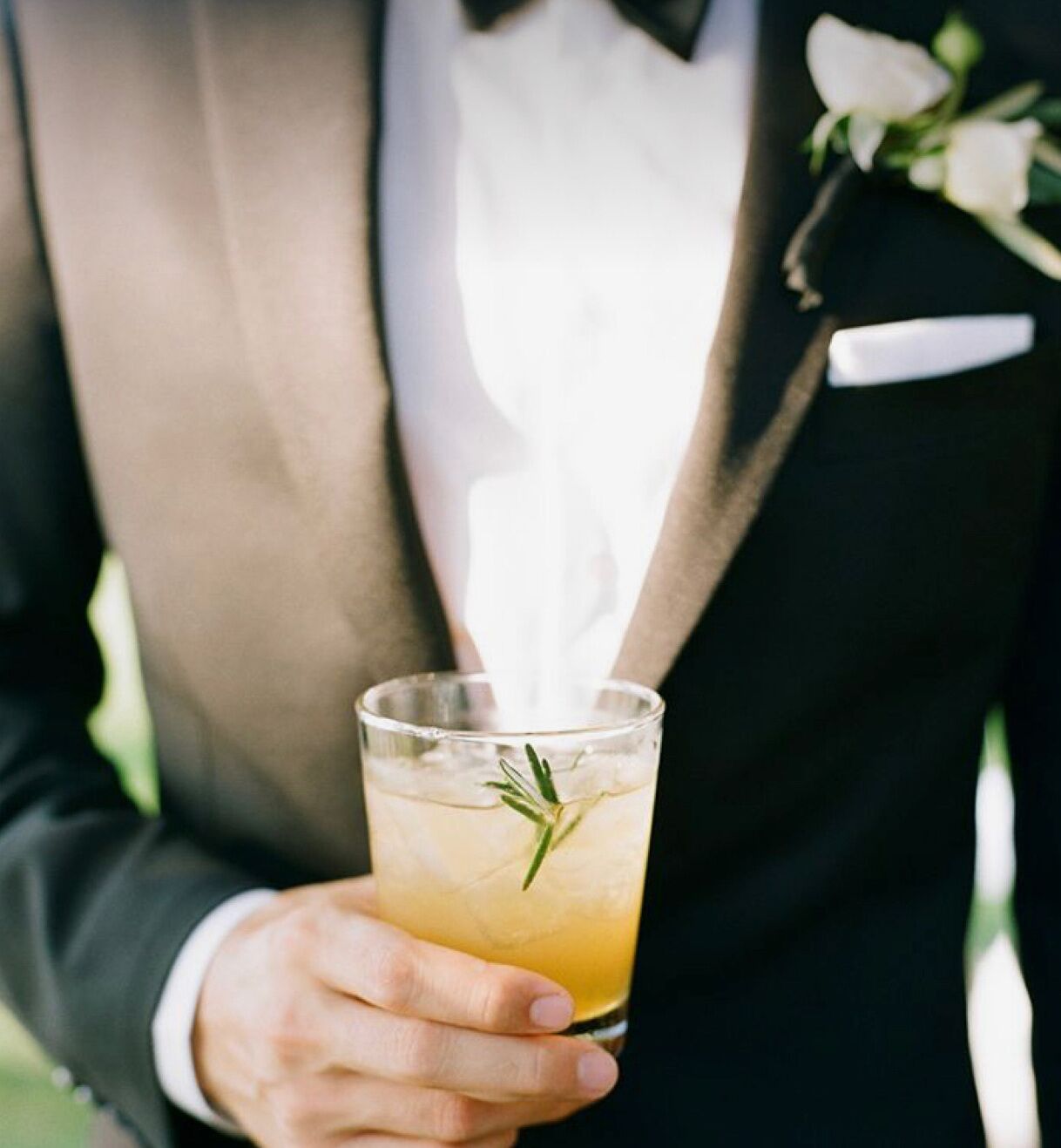 A person holding a glass with a drink and a sprig of rosemary.