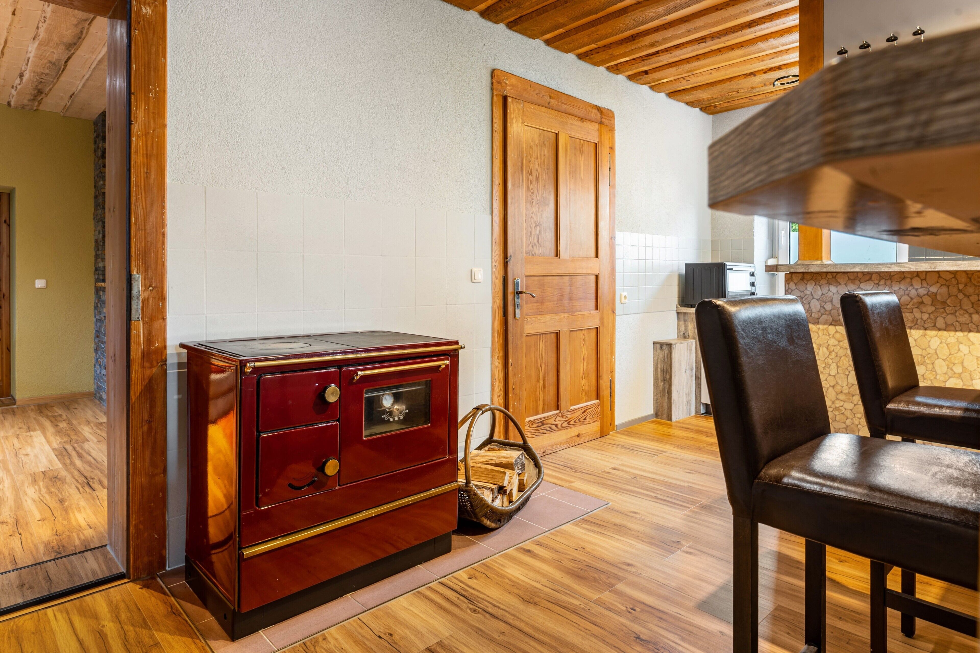 Une table de salle à manger en bois avec des chaises, une commode en bois rouge, une porte en bois et un coin cuisine doté d’un plan de travail en pierre.