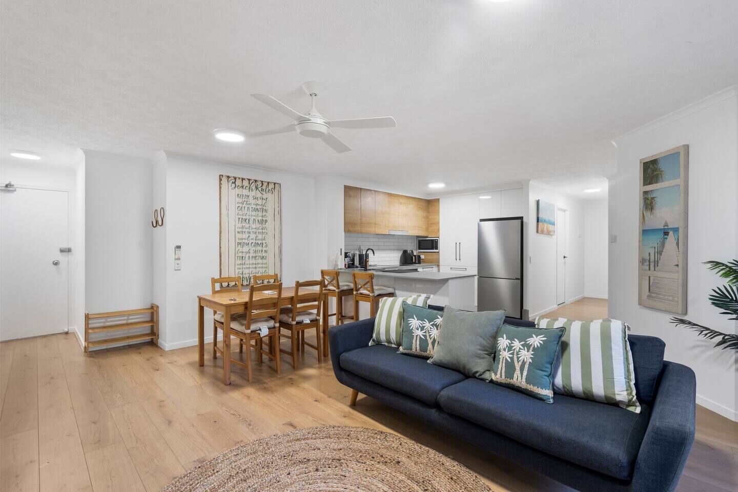 A modern living room with a sofa, a dining table with chairs, a kitchen area, and a ceiling fan.