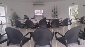 Un salón de conferencias con una mesa larga, sillas negras, una pantalla de televisión, banderas y un equipo de aire acondicionado.