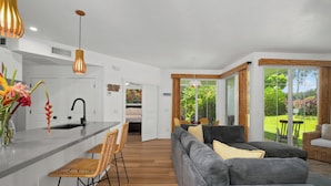 A modern kitchen with a central island, a dining area with a sofa and chairs, and a view of a garden through large windows. - Modern 2br Princeville Condo, Pool, Hot Tub, Fitness Center (Princeville)