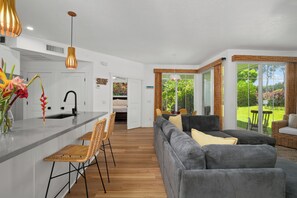 A modern kitchen with a central island, a dining area with a sofa and chairs, and a view of a garden through large windows. - Modern 2br Princeville Condo, Pool, Hot Tub, Fitness Center (Princeville)