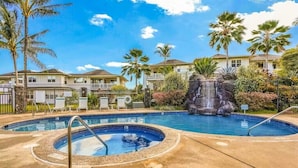 A swimming pool with a waterfall feature, surrounded by palm trees and a building with a balcony. - Modern 2br Princeville Condo, Pool, Hot Tub, Fitness Center (Princeville)