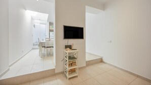 A modern, minimalistic room with a flat-screen TV mounted on the wall, a small white shelving unit with books, and a dining area visible in the background. - Holiday Home 'Lanzarote' with Private Terrace, Wi-Fi and Air Conditioning (Alvor)