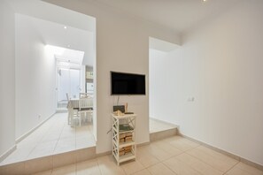 Une chambre moderne et minimaliste, avec un téléviseur à écran plat fixé au mur, une petite étagère blanche contenant des livres et un coin repas visible à l’arrière-plan.