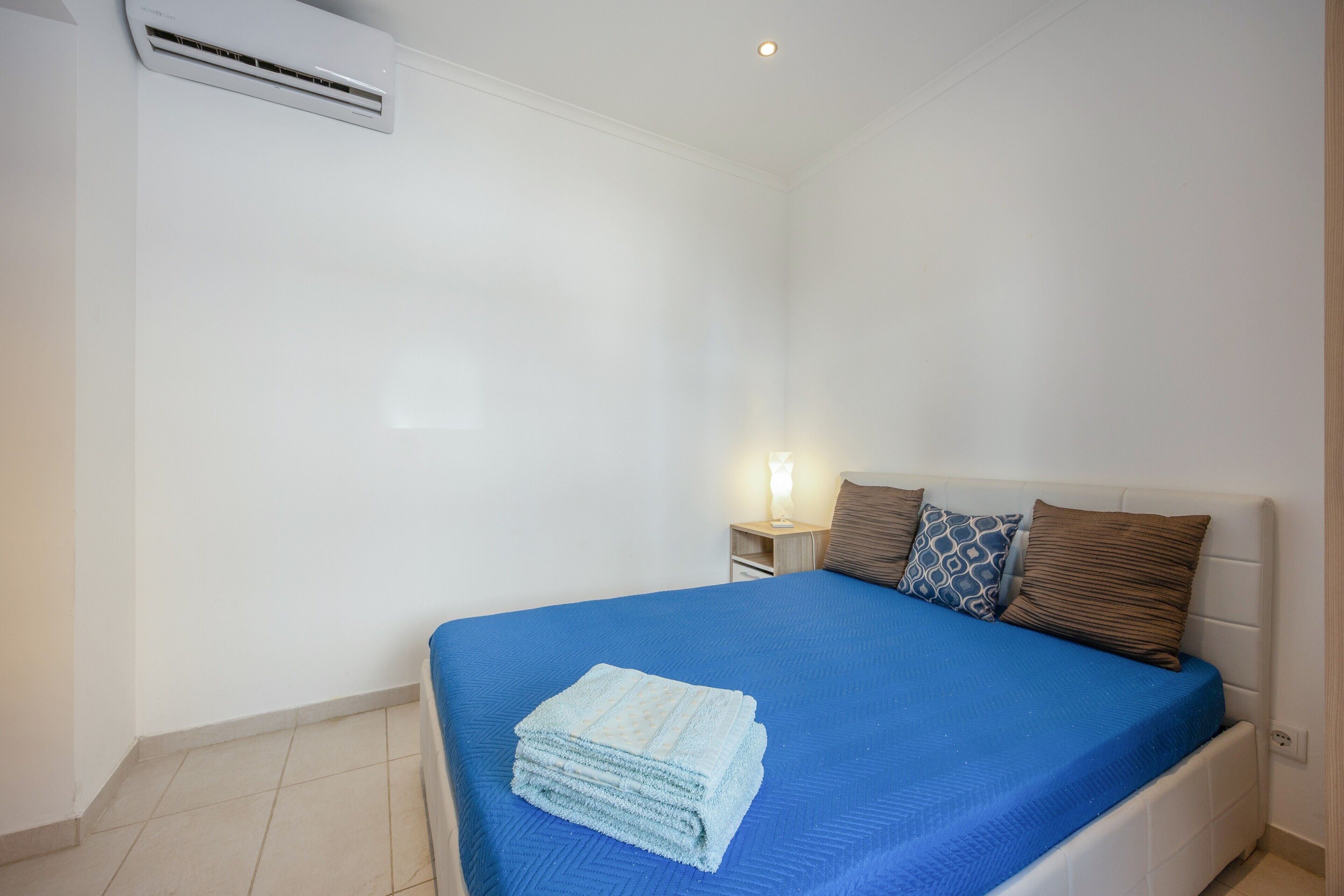 A hotel room with a bed, a nightstand, a lamp, and an air conditioning unit.