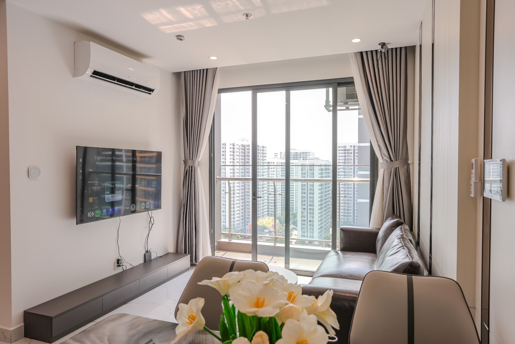 A modern living room with a sofa, a flat-screen TV mounted on the wall, and a large window with curtains.