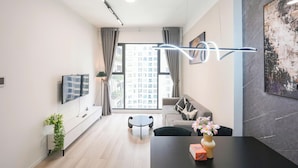 A modern living room with a sofa, a dining table, a TV mounted on the wall, and a large window with curtains. - Magnolia Vinhomes Apartment (Ho Chi Minh City)