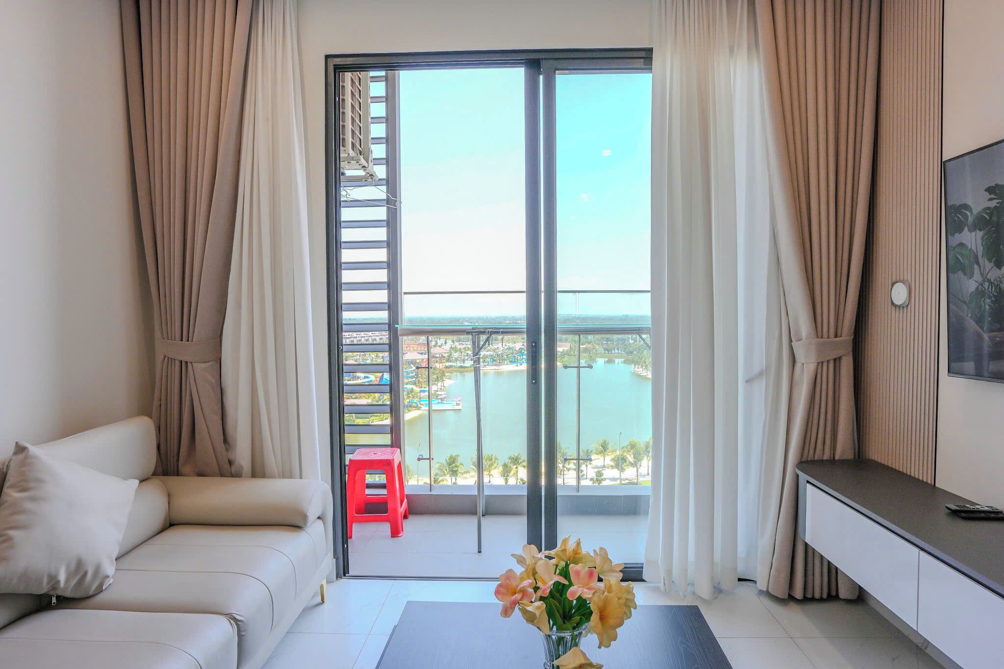 A room with a sofa, a small table with a vase of flowers, a red stool, and a view of a body of water through a glass door.