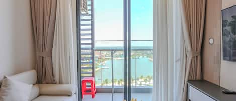 A room with a sofa, a small table with a vase of flowers, a red stool, and a view of a body of water through a glass door.