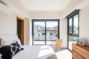 Ein Schlafzimmer mit einem großen Bett, einem Sessel, einer Kommode und einer Glastür zum Balkon mit Blick auf die Stadt.