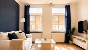 A modern living room with a sofa, coffee table, TV, and windows with curtains. - Maritime old town apartment Wismar with WLAN and workstation - 2 rooms (Wismar)